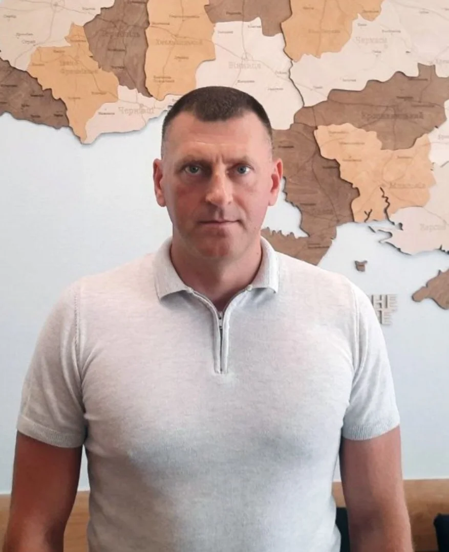Чоловік у світлому поло стоїть перед дерев’яною картою України
