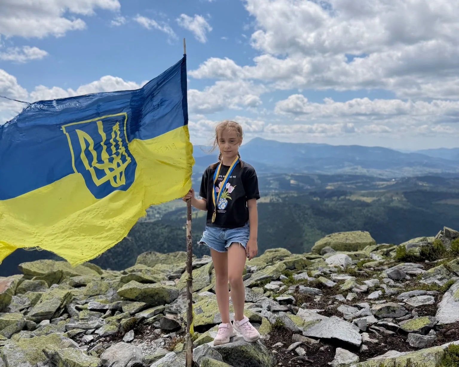 Дівчинка з медаллю тримає прапор України на скелястій вершині