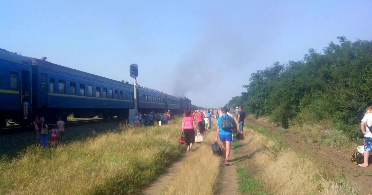 Пасажири йдуть стежкою біля синього потяга з димом попереду