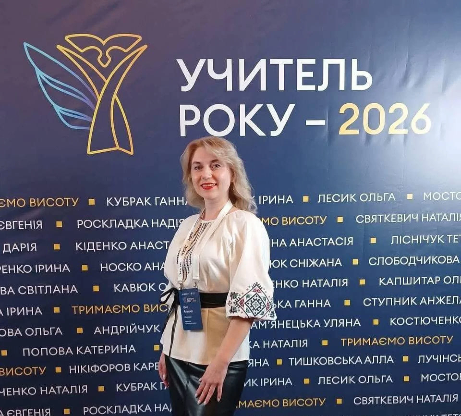 Жінка у вишиванці стоїть перед банером конкурсу Учитель року 2026