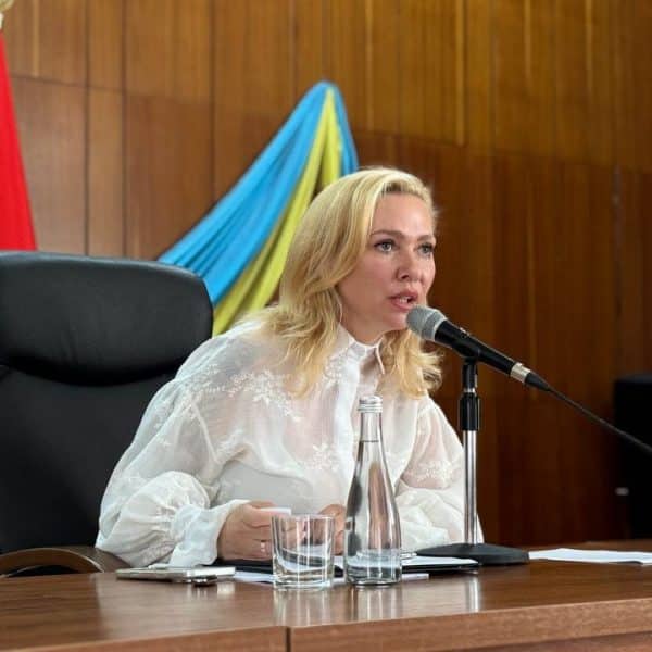 Білява жінка в білій блузці говорить у мікрофон за столом на тлі прапора України