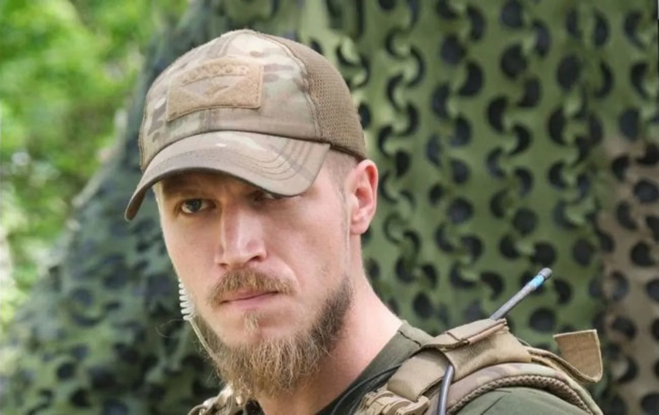 Військовий у камуфляжній кепці з нашивкою та тактичному спорядженні на тлі сітки