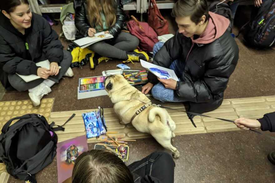 Молоді люди сидять на підлозі зі скетчбуками та фарбами поруч із мопсом